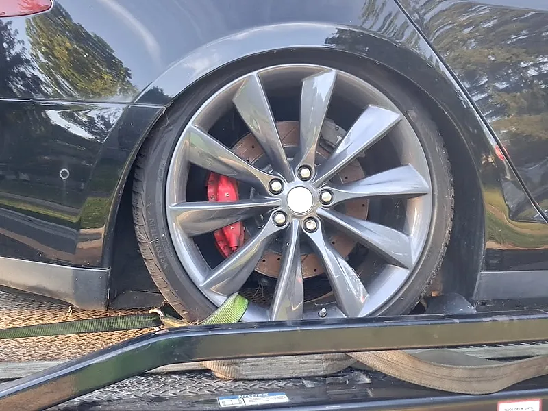 Close-up of a soft wheel strap wrapped around the rear tire of a black Tesla on a flatbed, with nothing hooked to the body or suspension
