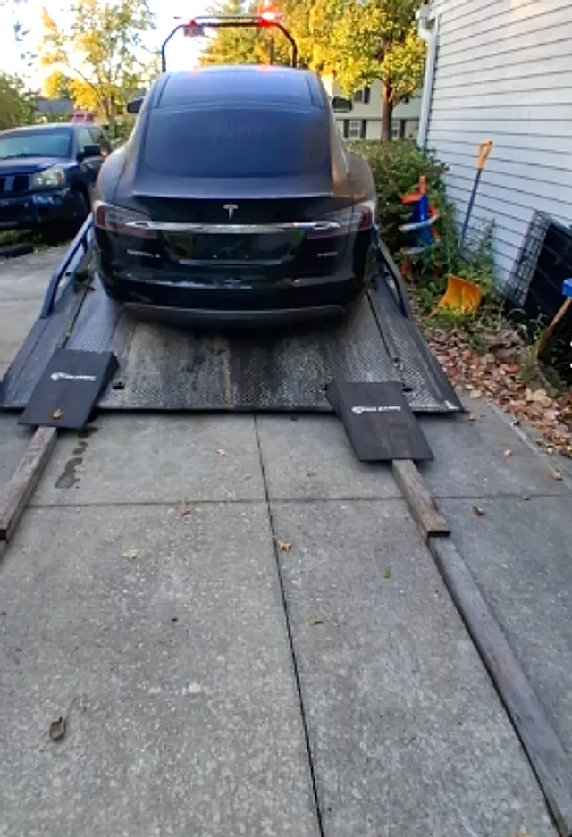 Rear view of a black Tesla Model S on a tilted flatbed, with two ramp pads and wooden boards placed at the tail of the deck